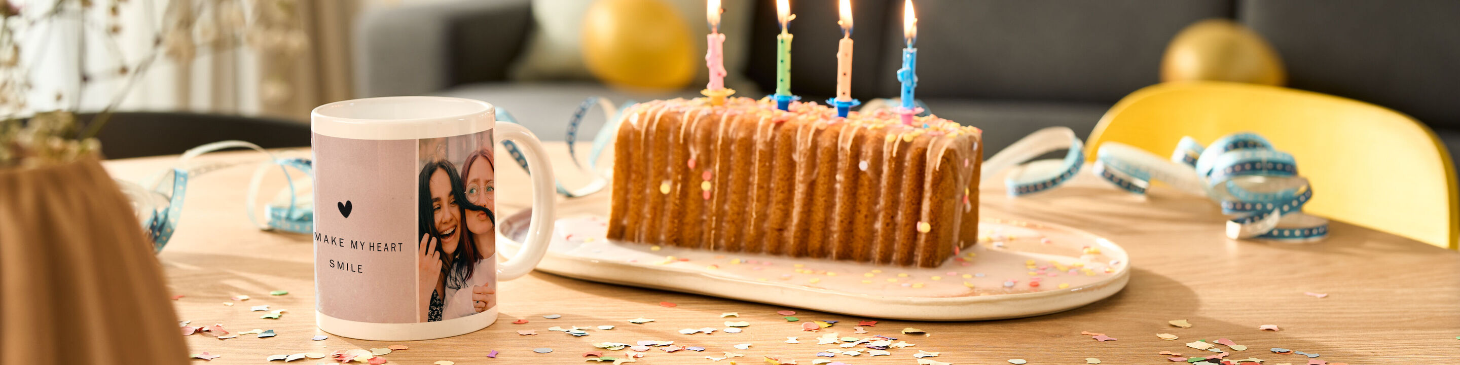 Una tazza di compleanno decorata con amore davanti a una torta colorata con candeline. Il tavolo è festosamente decorato.