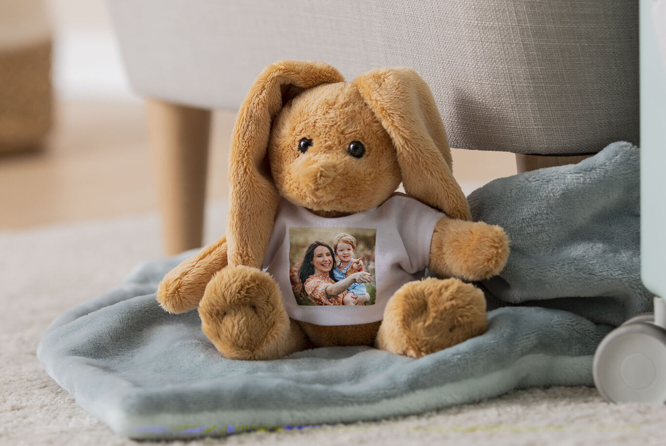 Un peluche marrone con lunghe orecchie seduto su un panno blu morbido. La maglietta mostra una famiglia felice.